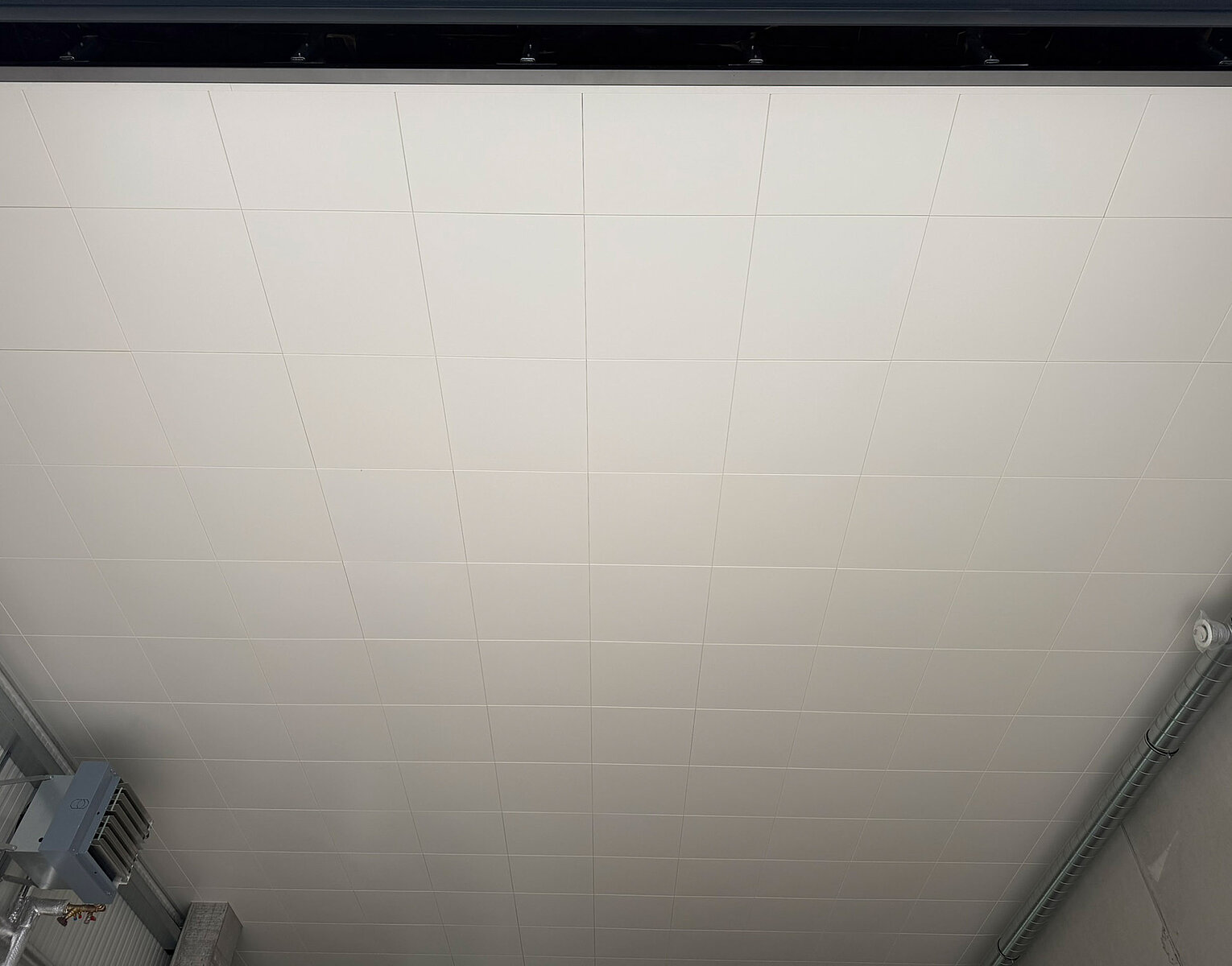 Weißer Deckenbereich mit quadratischen Paneelen in einem industriellen Innenraum mit sichtbaren Rohren an den Seitenwänden.
