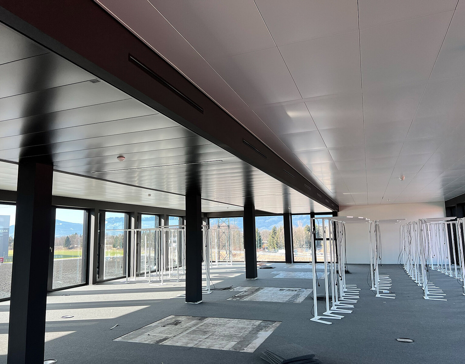 Großer leerer Raum mit vielen weißen Metallgestellen, großen Fenstern und schwarzer Decke mit Säulen, hellem Boden und Ausblick auf Außenbereich.