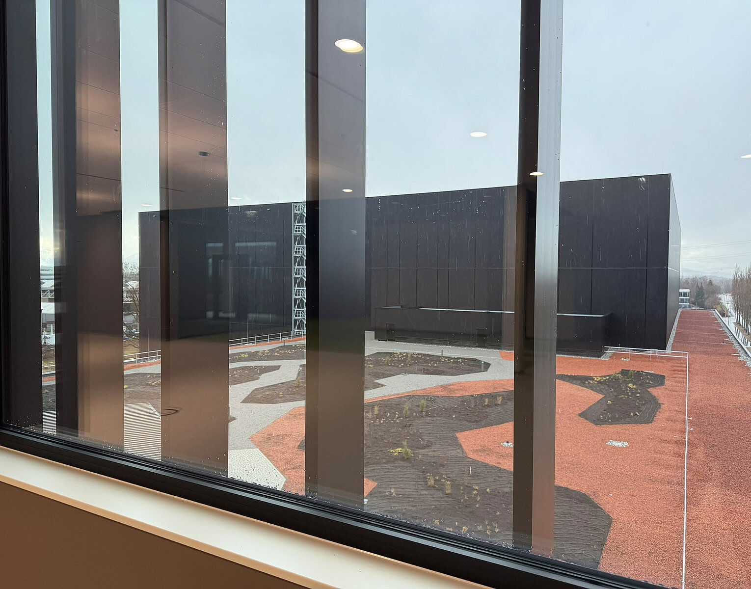 Blick durch eine Fensterfront auf ein modernes, schwarzes Gebäude mit niedrigem, flachem Dach und einem umliegenden Gartenbereich mit rotbrauner und schwarzer Bodenfläche.