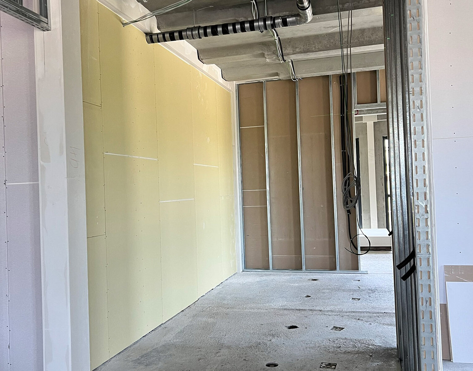 Raum im Rohbau mit sichtbaren Baustahlträgern und teils verputzten Wänden, Decke mit offenliegenden Rohrleitungen und Kabeln, Boden aus unbehandeltem Beton.