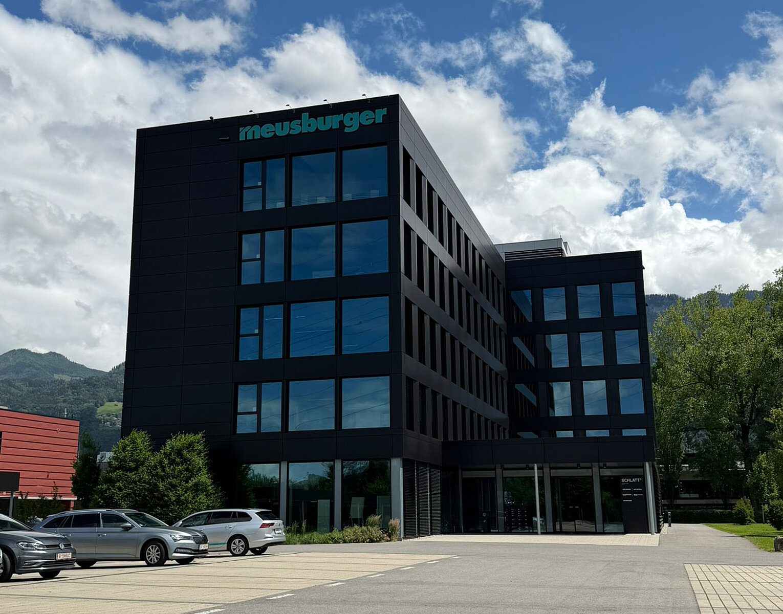 Modernes Bürogebäude mit schwarzer Fassade und großen Fenstern unter blauem Himmel mit Wolken, Parkplatz mit Autos im Vordergrund.