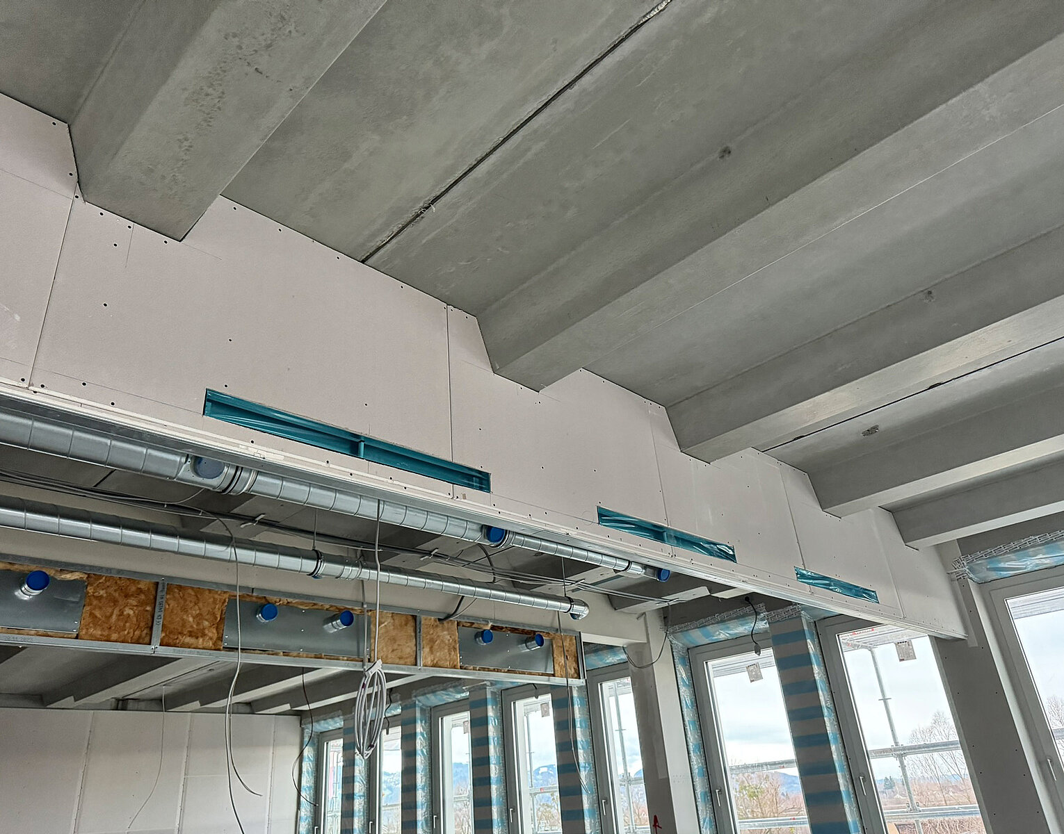 Innenraum einer Baustelle mit sichtbaren Betondecken, Lüftungsrohren und großen Fenstern, in denen teilweise eine Stadtansicht zu sehen ist.