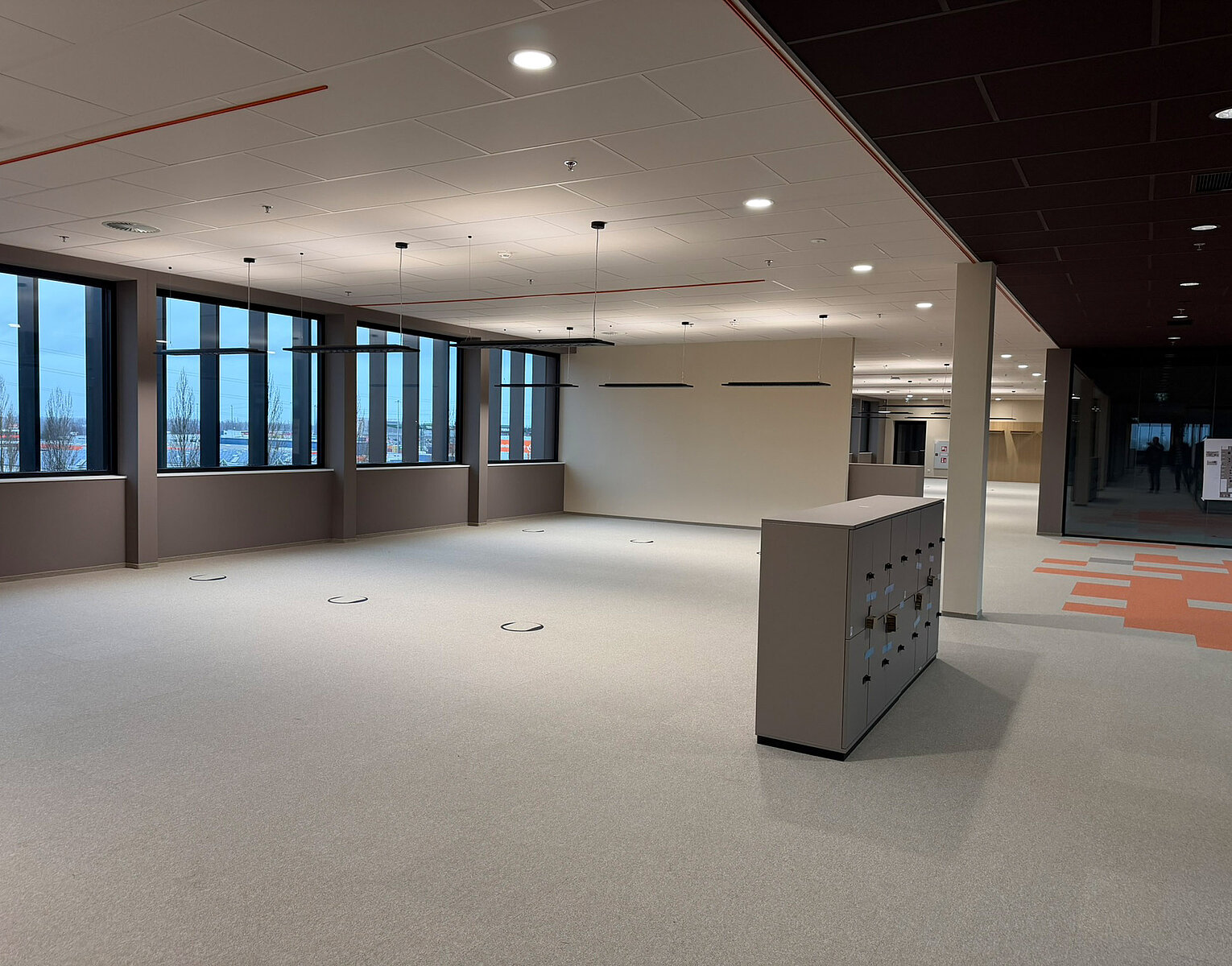 Leerer, moderner Büroraum mit großen Fenstern und hellgrahem Teppichboden, beleuchtet durch Deckenlampen, mit einem Schrank in der Mitte und Blick auf weitere Räume im Hintergrund.