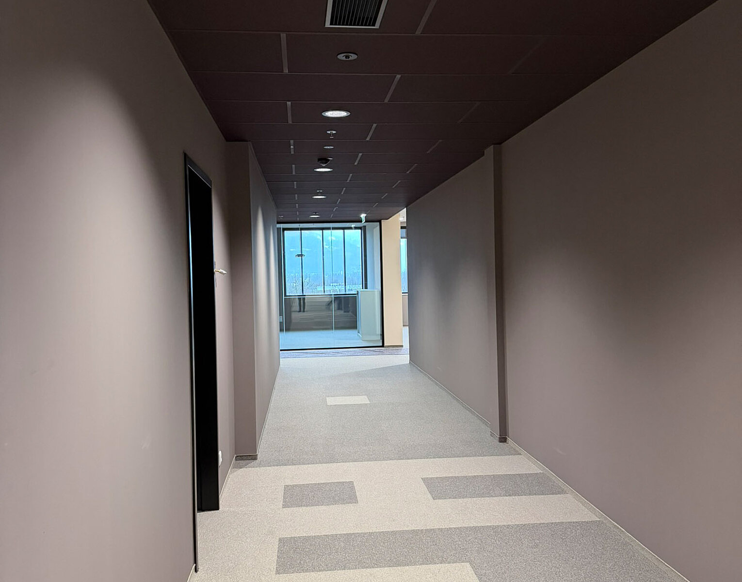 Blick in einen langen, schmalen Flur mit grauem Teppichboden, grauen Wänden und dunkler Decke, am Ende Fenster mit Blick nach draußen.