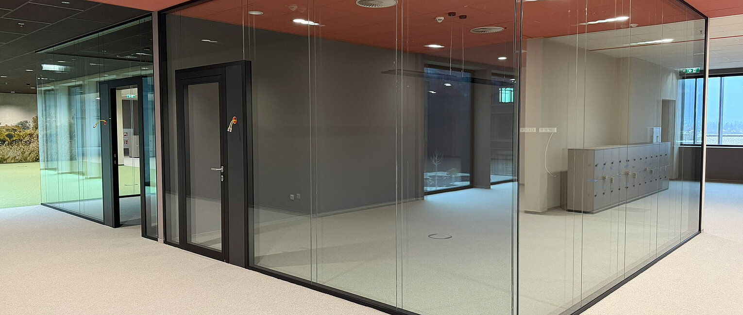 Moderne Büroeinheit mit großer Glas-Innenwand und glatter Teppichbodenfläche in hellen und dunklen Farbtönen, roter Decke und integrierten Deckenleuchten.