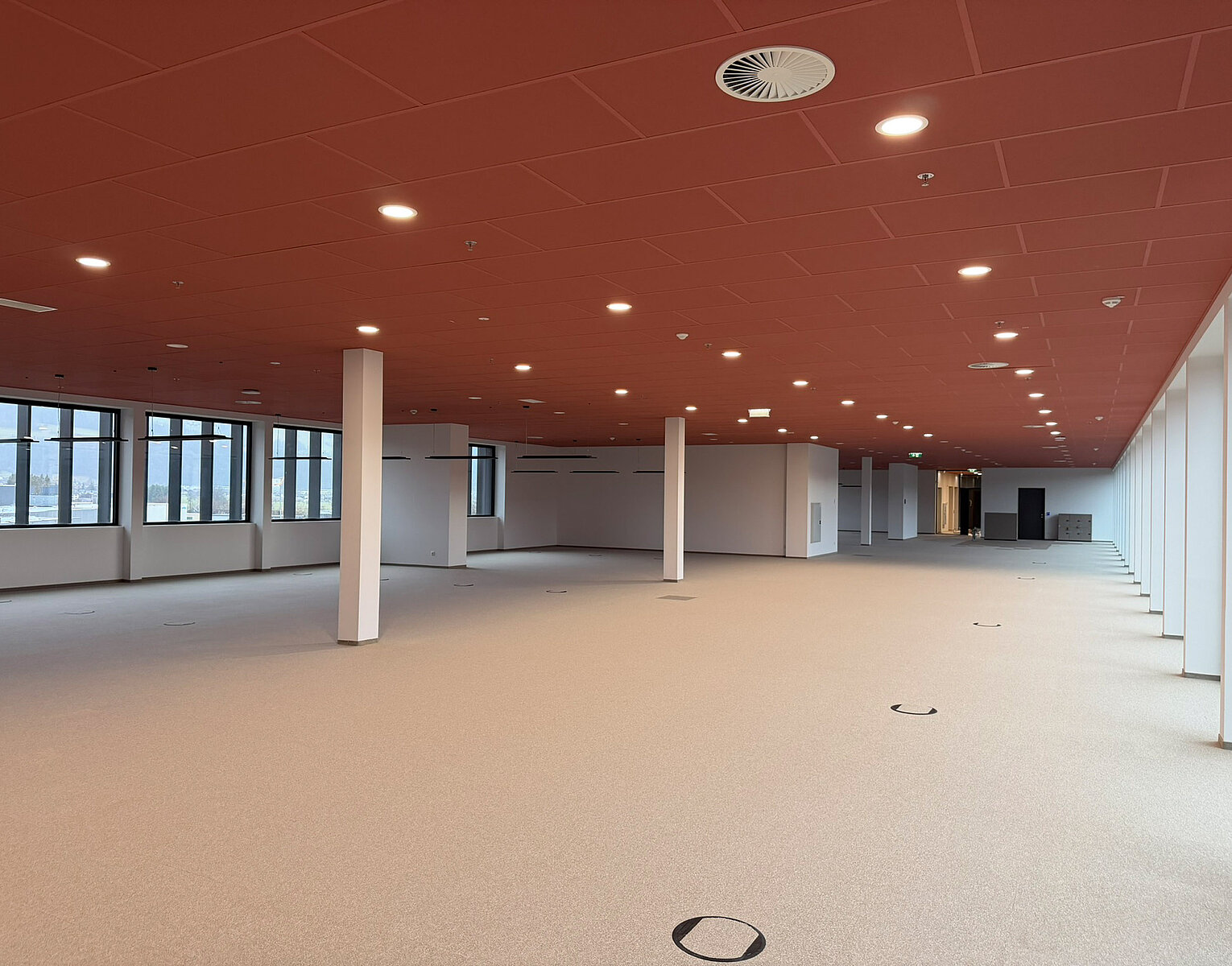 Großer, leerer Büroraum mit roter Decke, weißen Stützsäulen und großen Fenstern entlang der Wände, der viel Tageslicht hereinlässt.