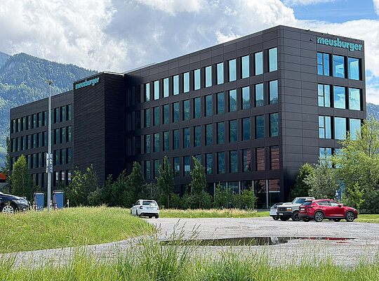 Modernes Bürogebäude mit mehreren Etagen und großen Fenstern, umgeben von Bäumen und Parkplätzen mit mehreren geparkten Autos vor bewölktem Himmel und Bergen im Hintergrund.