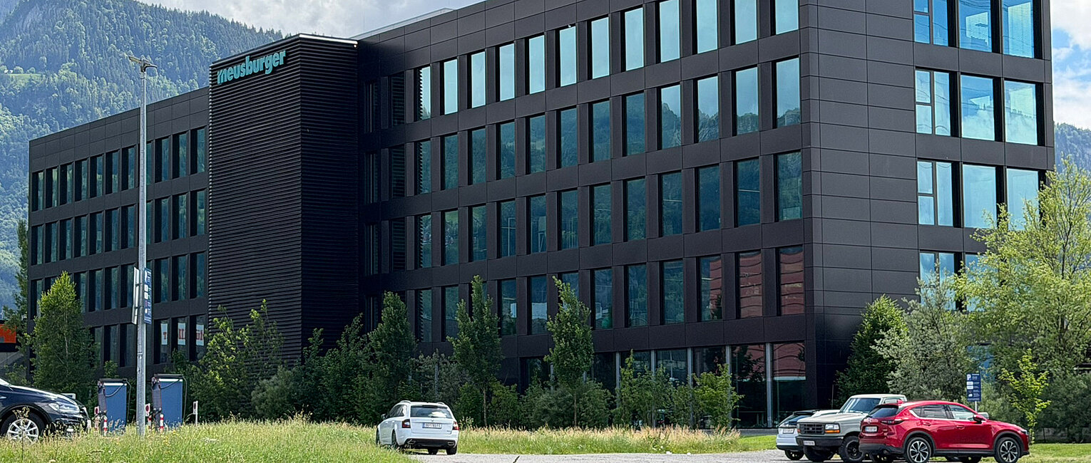 Modernes Bürogebäude mit mehreren Etagen und großen Fenstern, umgeben von Bäumen und Parkplätzen mit mehreren geparkten Autos vor bewölktem Himmel und Bergen im Hintergrund.