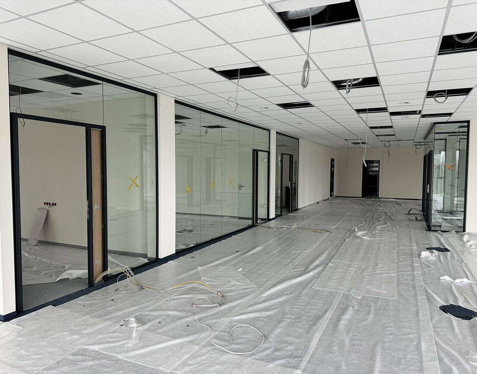 Großer Büroraum im Rohbau mit abgehängter Decke, Glaswänden und provisorisch abgedechtem Boden, Kabeln liegen lose herum, Raum ist unmöbliert und wird renoviert.