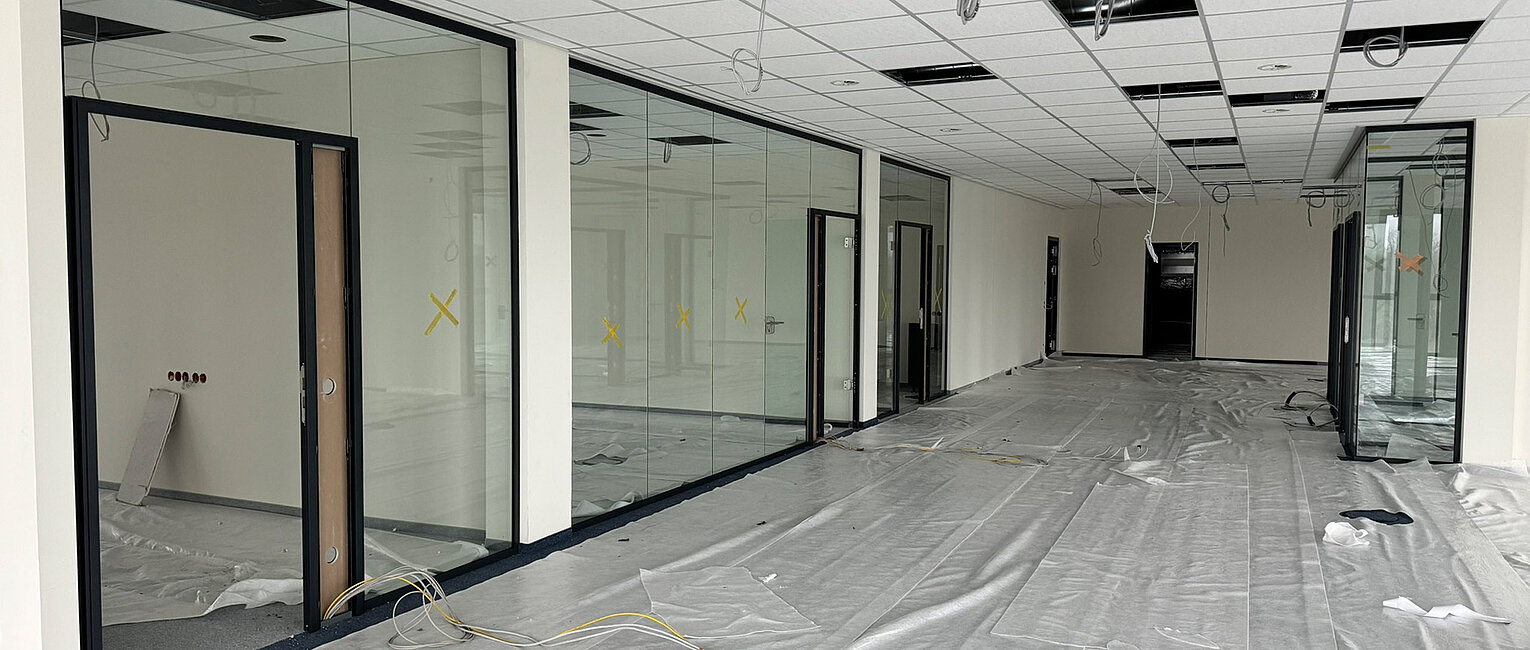 Großer Büroraum im Rohbau mit abgehängter Decke, Glaswänden und provisorisch abgedechtem Boden, Kabeln liegen lose herum, Raum ist unmöbliert und wird renoviert.