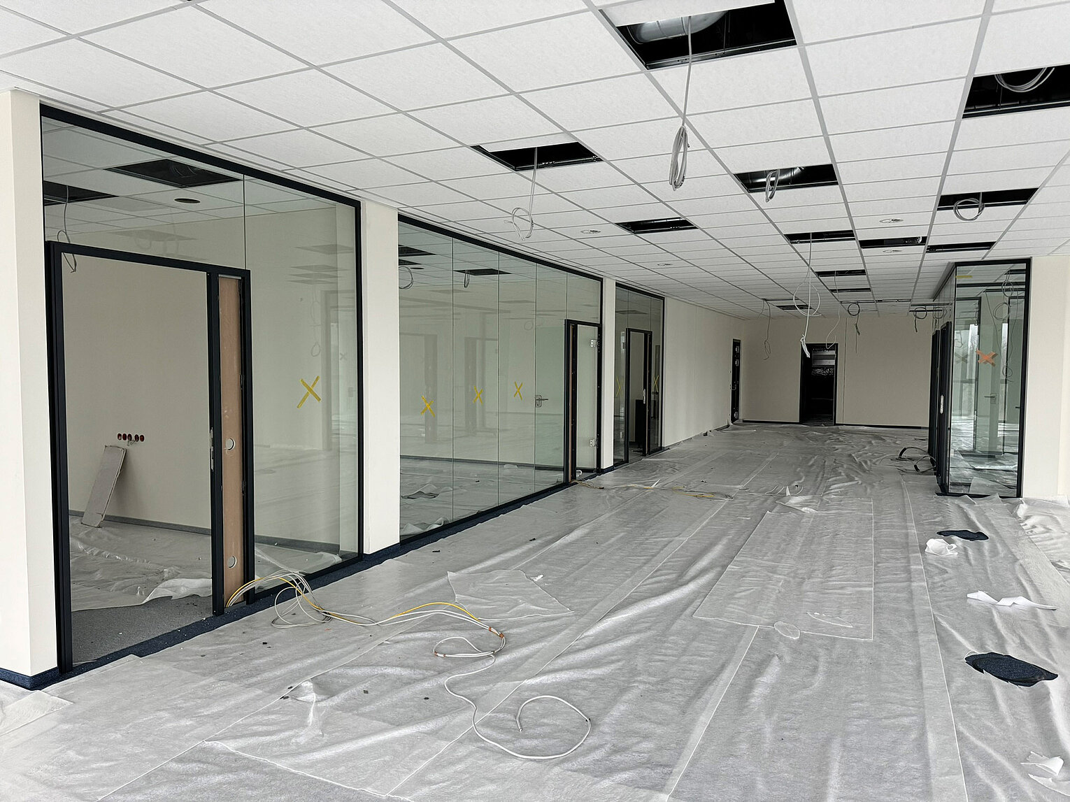 Großer Büroraum im Rohbau mit abgehängter Decke, Glaswänden und provisorisch abgedechtem Boden, Kabeln liegen lose herum, Raum ist unmöbliert und wird renoviert.