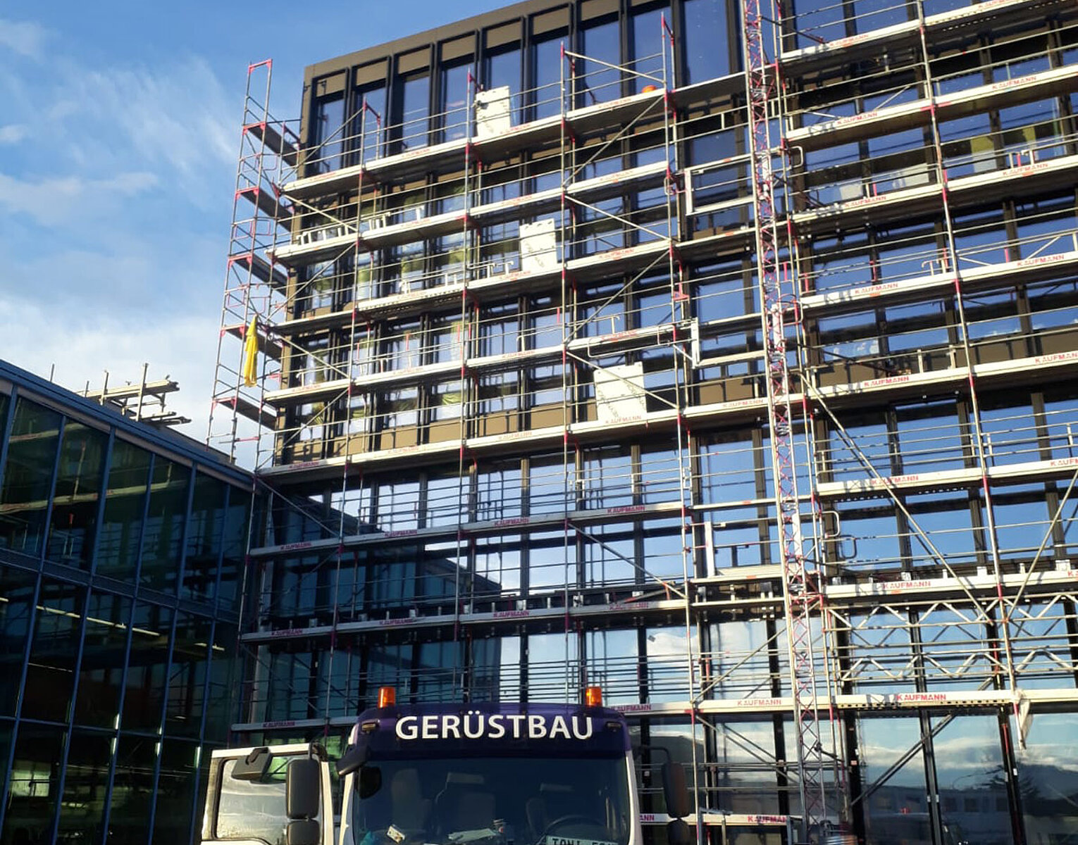 Fassadengerüst an einem mehrstöckigen Gebäude mit Baustellenfahrzeug und Arbeiter im Vordergrund bei klarem Himmel.
