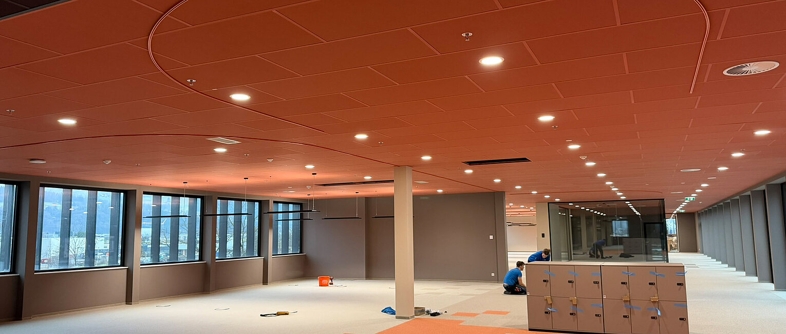 Großer leerer Büroraum mit orangen und grauen Teppichfliesen, oranger Decke mit runden Ausschnitten und großen Fenstern auf einer Seite, zwei Personen bei Arbeiten auf dem Boden.