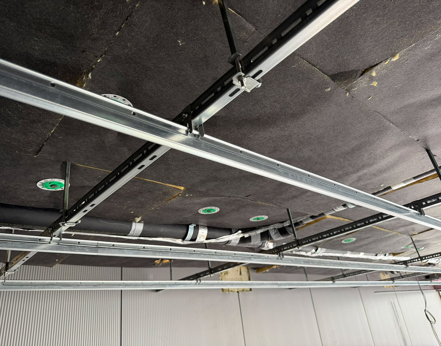 Decke mit installierten Metallträgern und Rohrleitungen sowie mehreren runden Einbauleuchten in schwarzer Dämmung.