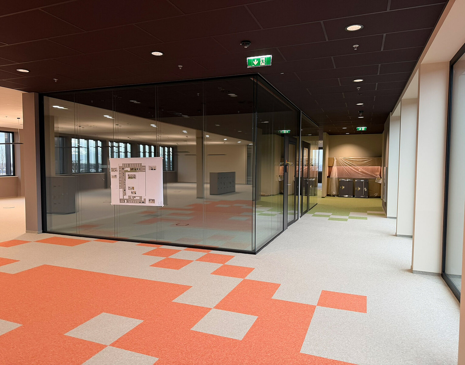 Moderner Büroraum mit großem Glasbüro, orangen und grauen Teppichfliesen und deckenhohen Fenstern, der eine helle und offene Arbeitsumgebung zeigt.