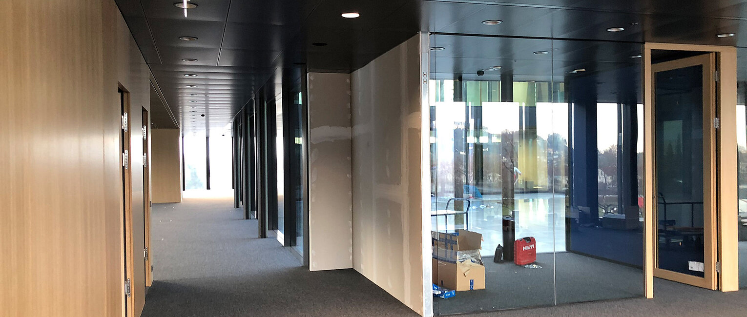 Moderner Büroflur mit Glaswänden und Holztüren, leer und gut beleuchtet, mit Kartons und Taschen im sichtbaren Raum hinter der Glaswand.