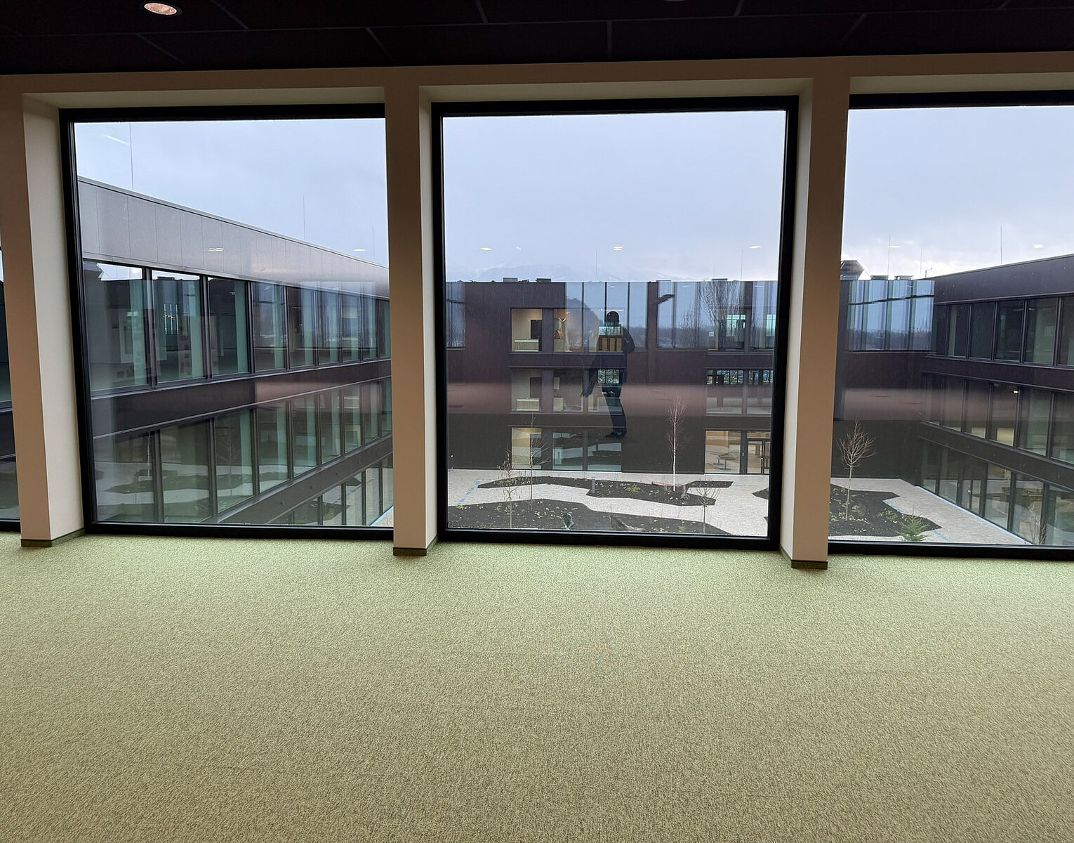 Blick durch drei große Fenster auf den Innenhof eines modernen Bürogebäudes mit Glasfassaden und grünem Teppichboden im Innenraum.