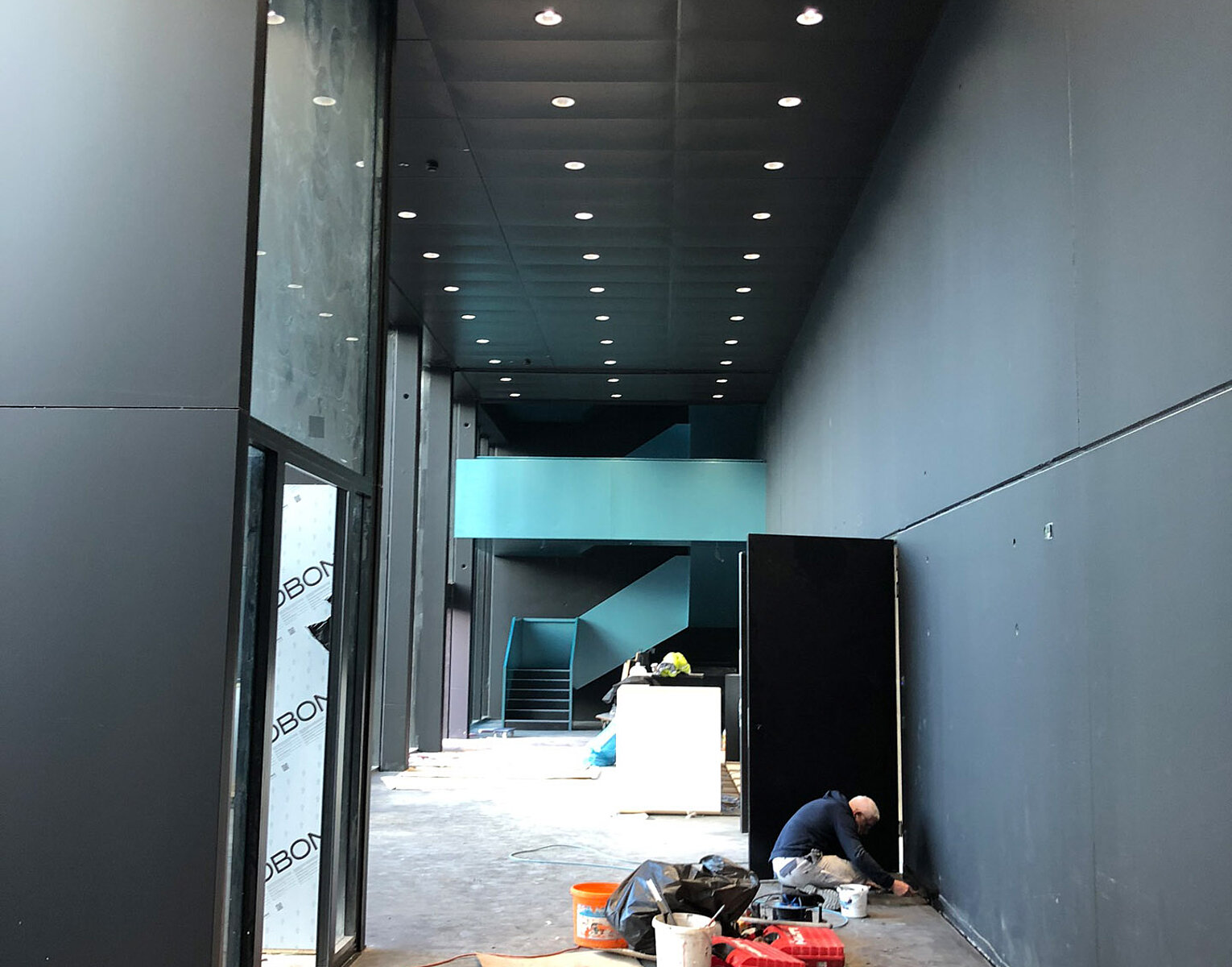 Innenraum eines modernen Gebäudes mit hoher Decke und schwarzen Wänden, Handwerker arbeitet auf dem Boden an der rechten Wand, verschiedene Werkzeuge und Baumaterialien liegen verstreut.
