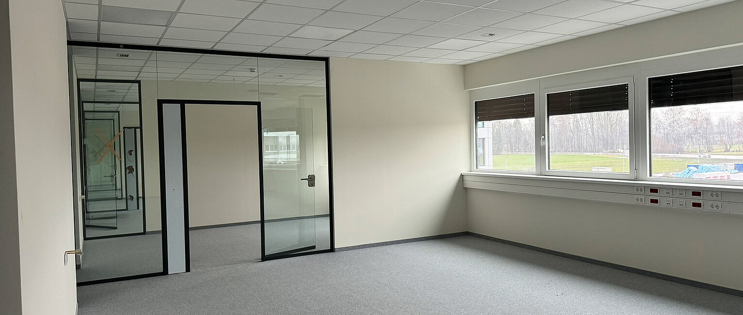 Leerer Büroraum mit grauem Teppichboden, weißen Wänden, großen Fenstern und einer Glaswand mit Tür. Im Hintergrund spiegelt sich der Raum mehrfach.