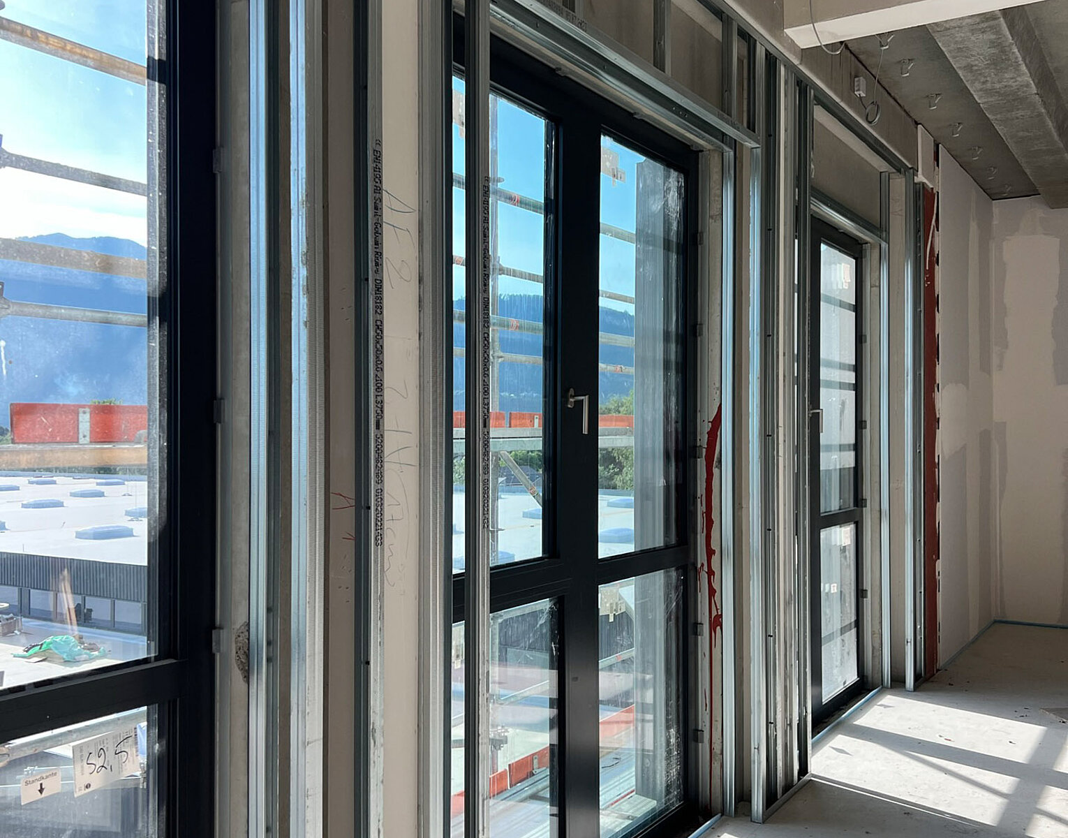 Blick auf große Fenster in einem unfertigen Raum mit sichtbaren Metallrahmen und Betonboden, durch die das Tageslicht einfällt. Draußen sind Industriegebäude und Container zu sehen.