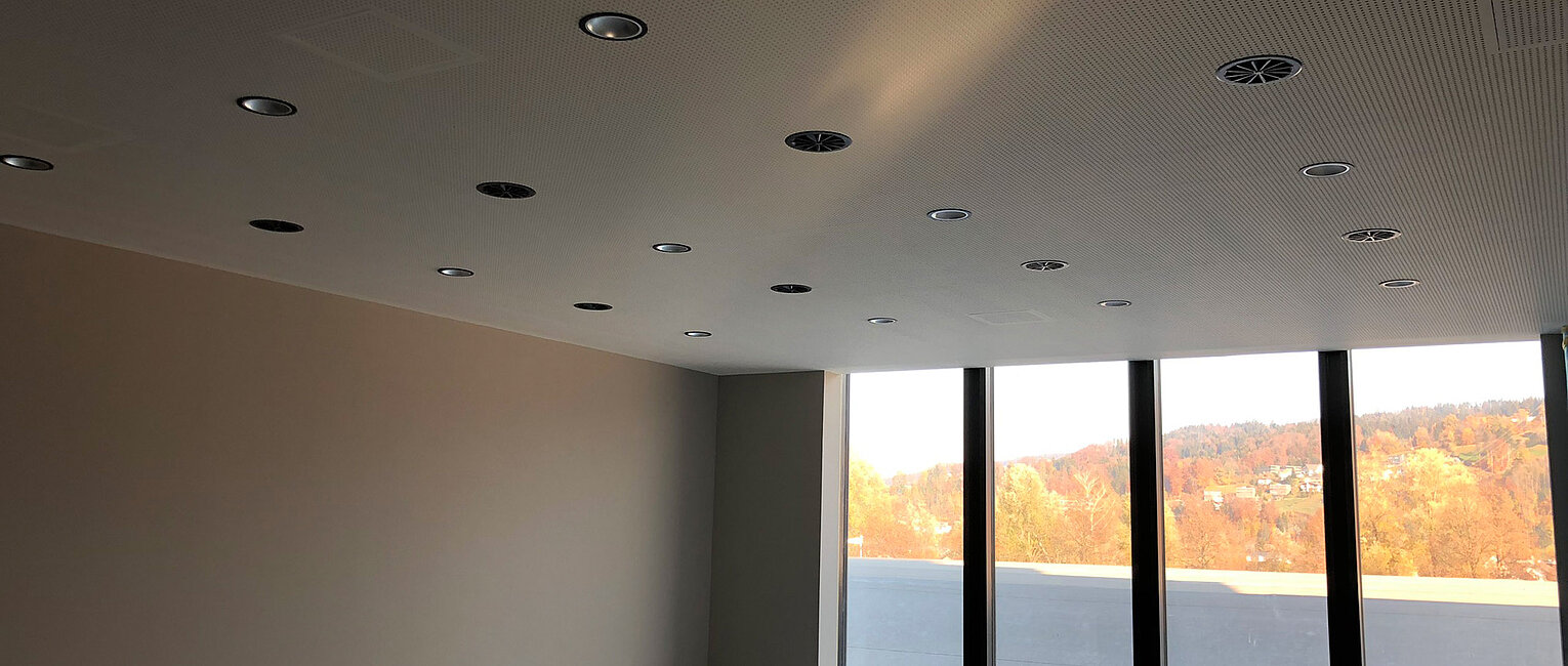 Leerer Raum mit hellgrauen Wänden, Decke mit vielen Einbaustrahlern und großen Fensterfronten, durch die herbstliche Bäume und ein sonniger Himmel zu sehen sind.