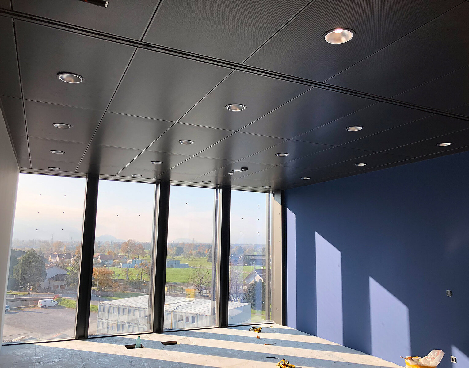 Innenraum eines modernen, leeren Raumes mit großer Fensterfront und Blick auf eine Landschaft mit Wiesen, Häusern und Bergen im Hintergrund. Sonneneinstrahlung erzeugt Lichtreflexe an einer dunkelblauen Wand.