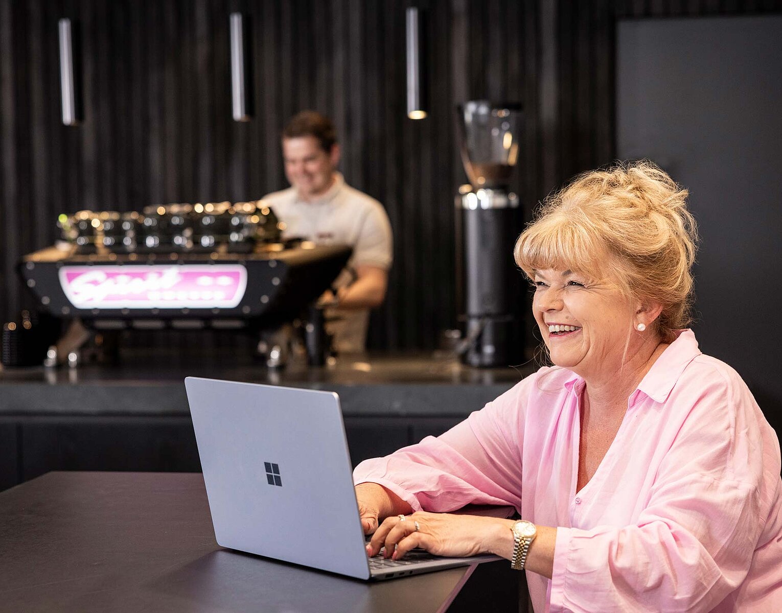 Ältere Frau sitzt lächelnd an einem Laptop in einem Café, während im Hintergrund ein Barista an der Espressomaschine arbeitet.
