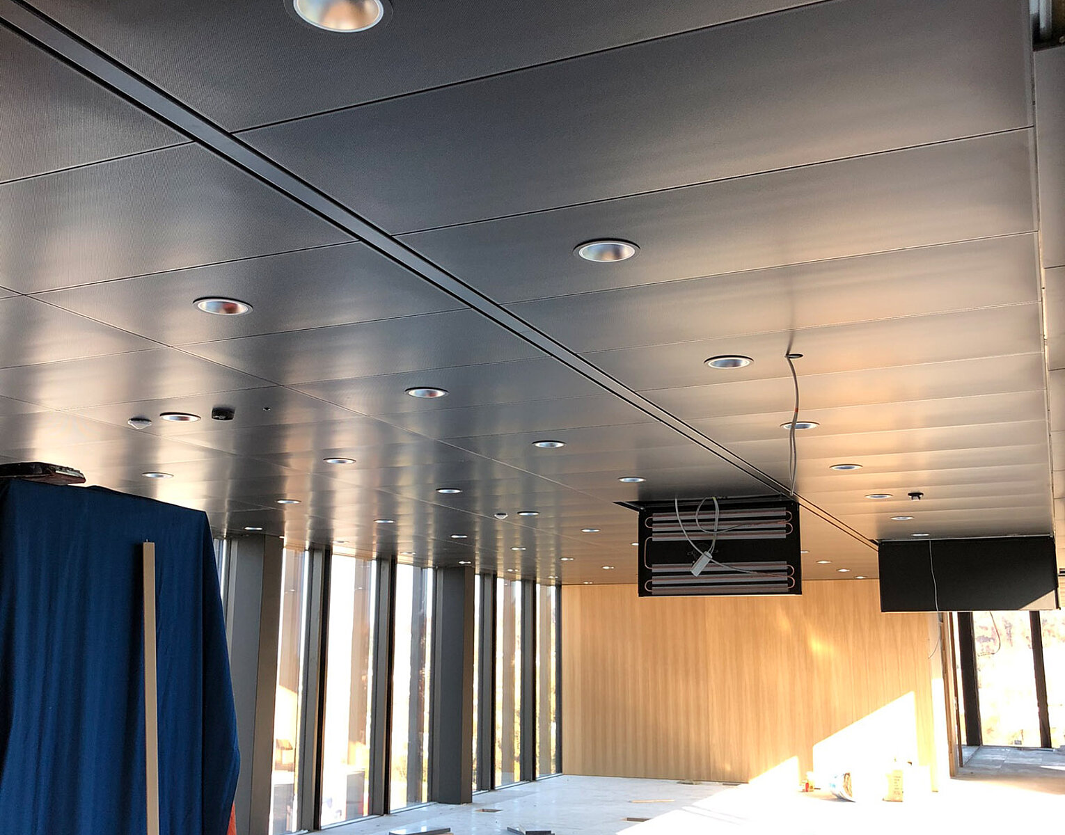 Großer leerer Raum mit bodentiefen Fenstern, Decke mit Einbauleuchten, unausgebauter Boden und offenem Kabelschacht, Sonnenlicht fällt durch Fenster.