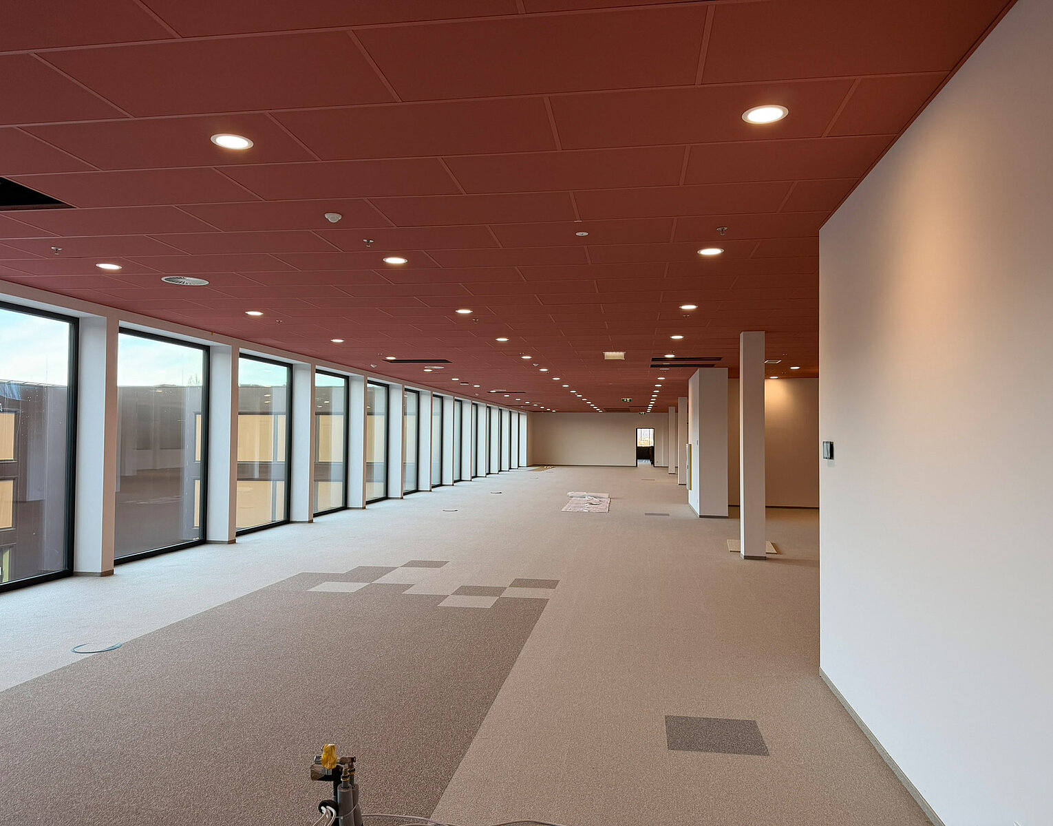 Großer leerer Büroraum mit roten Deckenplatten, grauem Teppichboden und bodentiefen Fenstern entlang einer Seite. Lichtquellen sind Deckenlampen. Einige weiße Säulen und eine weiße Wand sind im Raum sichtbar.