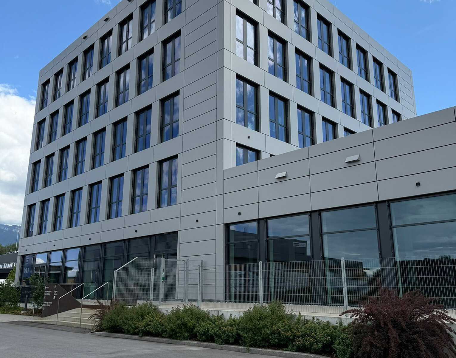 Moderner mehrstöckiger Bürogebäude mit vielen Fenstern, klarer Himmel mit wenigen Wolken im Hintergrund, umgeben von Gehweg und kleinen Sträuchern.