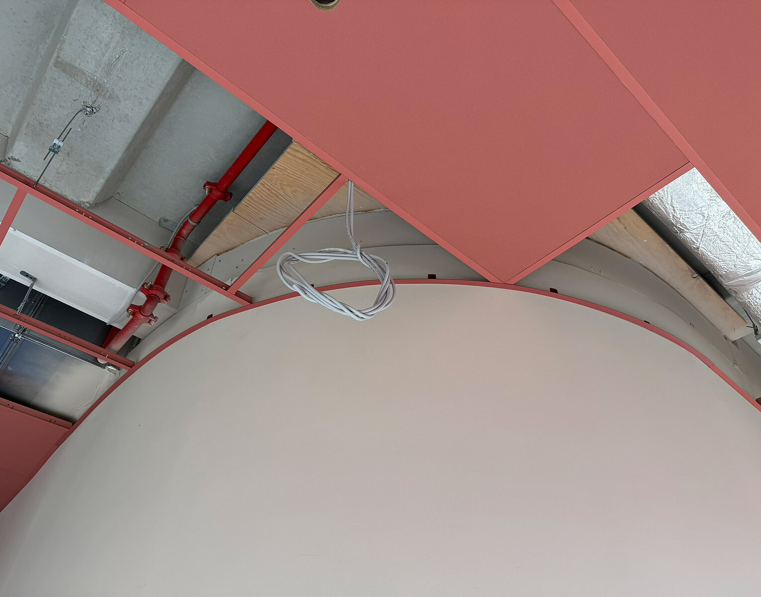 Blick an eine Decke mit rotem Gestell, roten Abdeckpaneelen und einem großen weißen, gebogenen Element, Kabel hängen lose von der Decke.