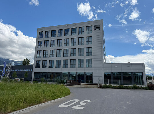Modernes mehrstöckiges Bürogebäude mit Glasfenstern, davor eine Straße mit Geschwindigkeitsbegrenzung 30, umgeben von Wiesen und blauem Himmel mit Wolken.