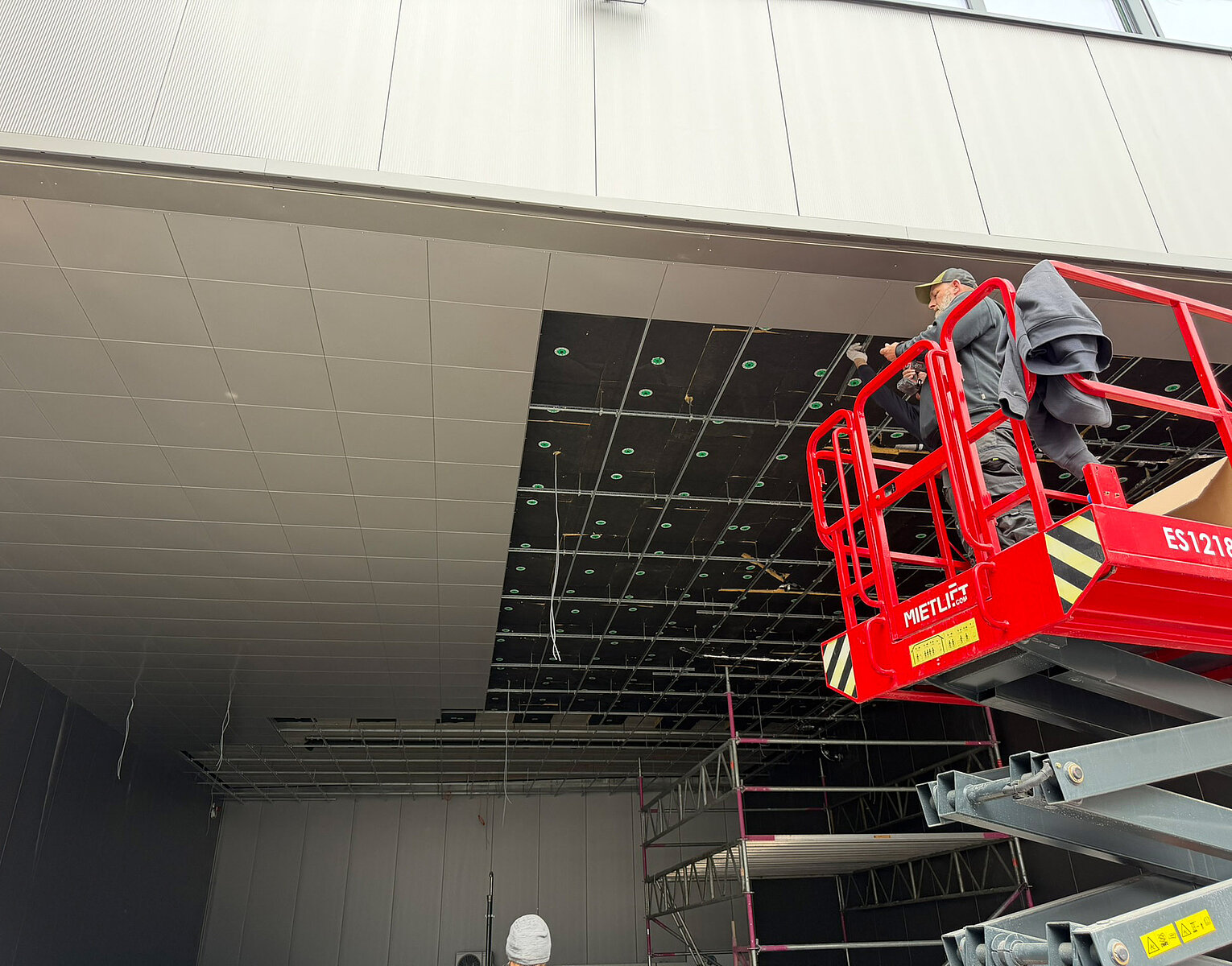Zwei Bauarbeiter arbeiten auf einer Hebebühne an der Installation oder Reparatur der Deckenverkleidung in einem großen Innenraum. Die Deckenplatten sind teilweise entfernt, um darunter liegende Leitungen und Strukturen freizulegen.