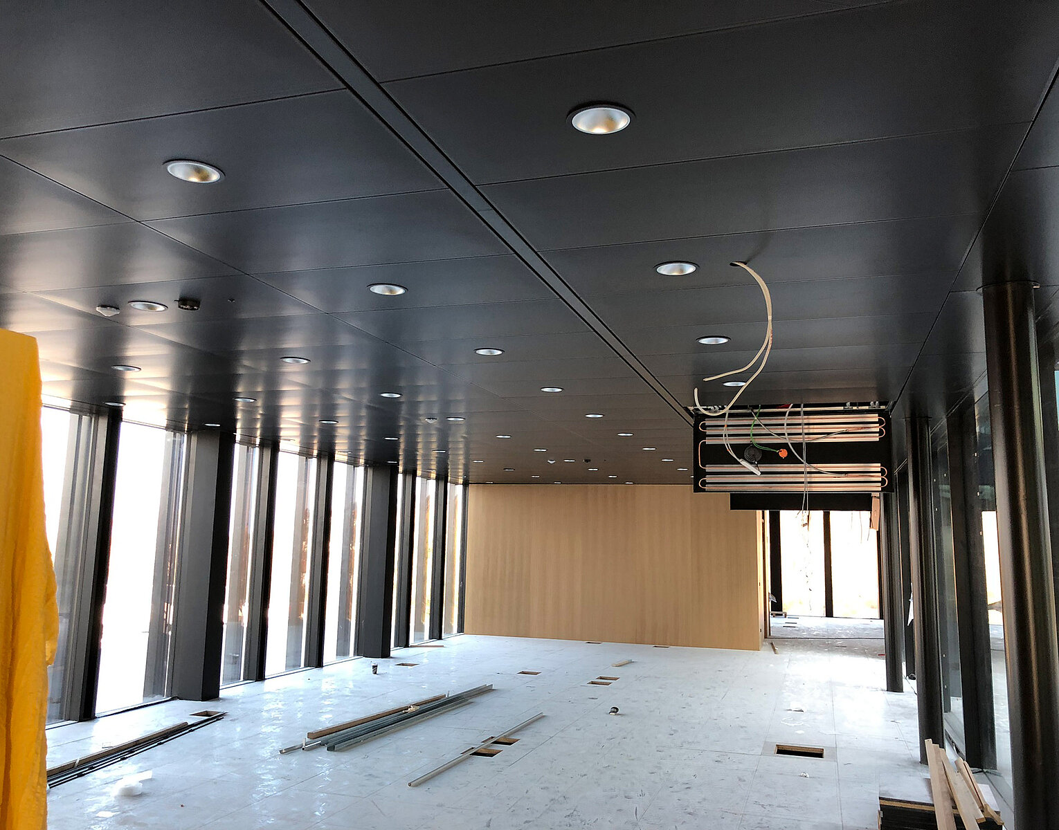 Innenansicht eines unmöblierten, modernen Büroraums mit großen Fenstern, dunkler Decke mit eingebauten Lampen und offenliegenden Kabeln, während Bauarbeiten im Gange sind.