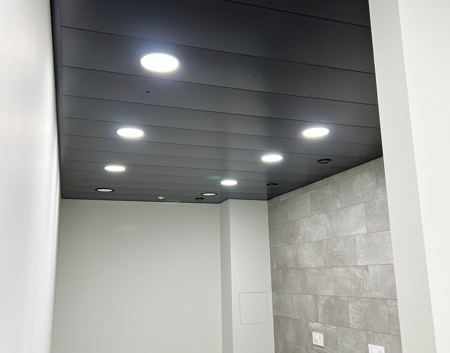 Schlichter Raum mit grauer Kachelwand, weißen Wänden, zwei Wandtoiletten und einer schwarzen Decke mit mehreren eingebauten, hellen Deckenleuchten.