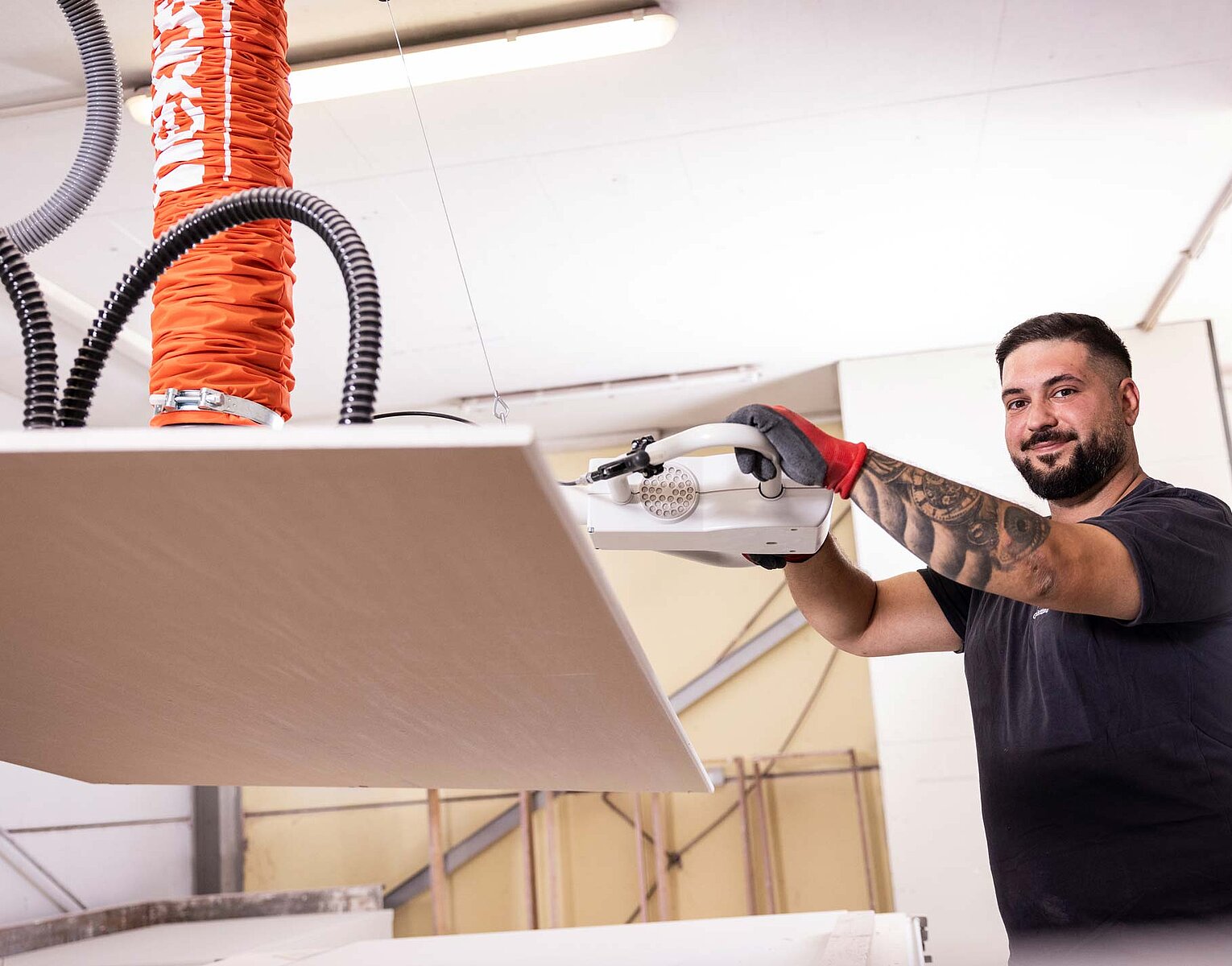 Ein Mann mit Tattoos und Handschuhen bedient in einer Werkstatt eine Hebemaschine, die eine große Platte anhebt.