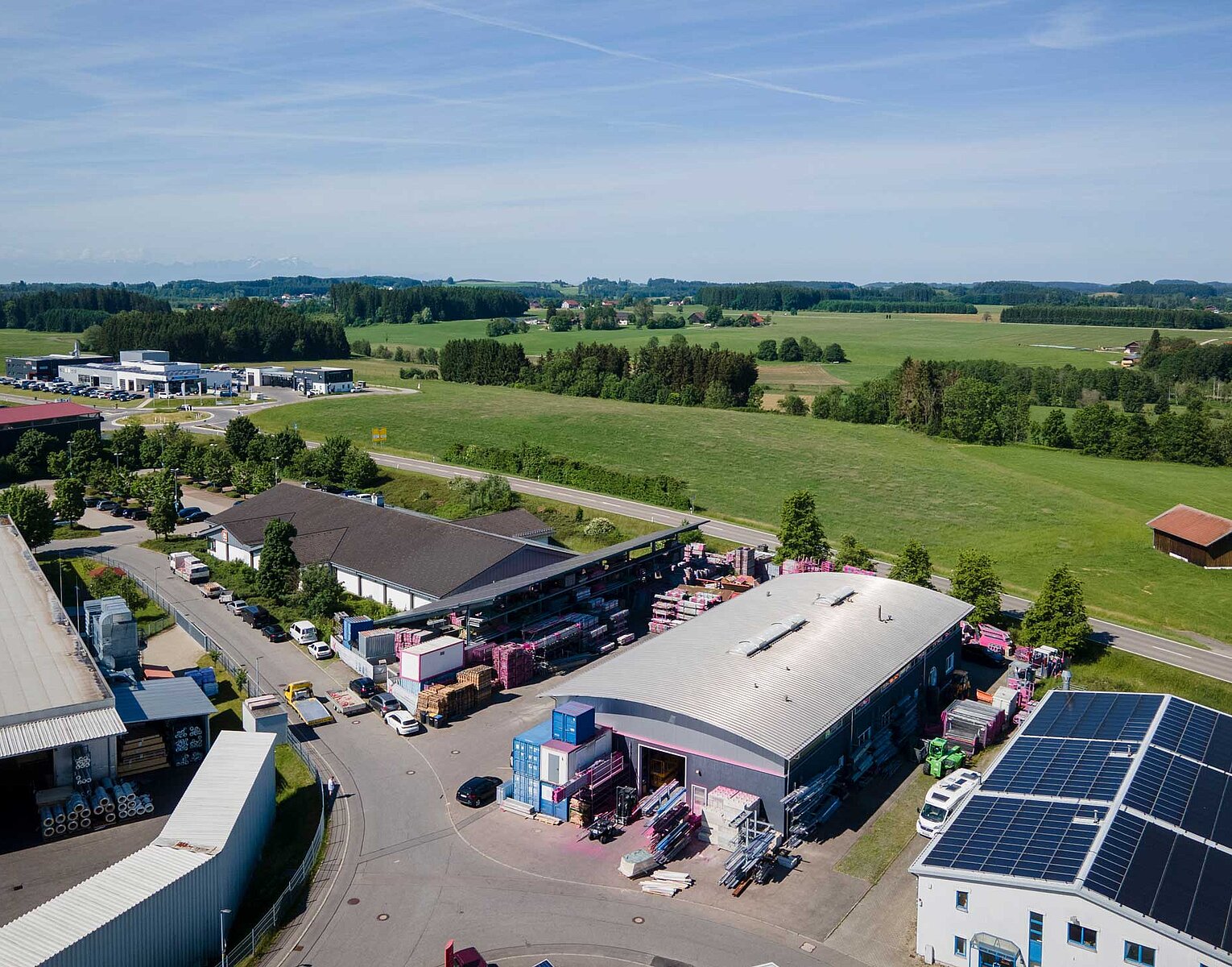 Luftaufnahme eines Industriegebiets mit mehreren Gebäuden, Parkplätzen, Lastwagen und grünen Feldern im Hintergrund unter blauem Himmel.