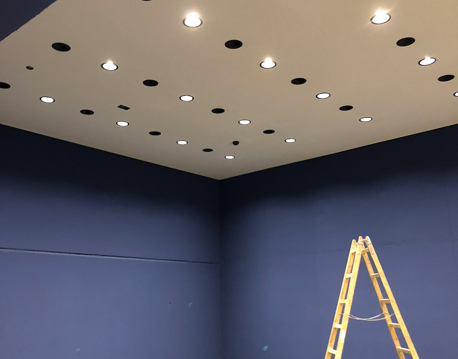 Raum mit blauen Wänden und Decke, an der mehrere Einbauleuchten installiert sind, daneben steht eine gelbe Leiter und auf dem Boden liegt ein Wurfhelm.