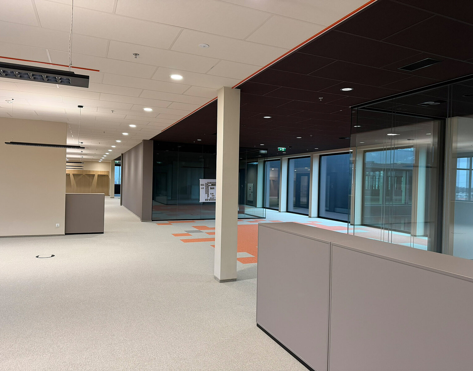 Leeres modernes Büro mit hellen Wänden, Glaswänden und Teppichboden, beleuchtet durch Deckenlampen.